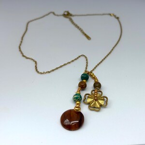 Op de afbeelding: Een goudkleurige ketting met een hanger met een klavertjevier en een cluster van kralen in bruin-, groen- en goudtinten. De ketting heeft ook een grote, ronde, bruine hanger.