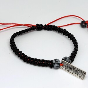 Op de afbeelding: Zwart gevlochten armband met rode accenten en verstelbare rode koorden. De armband heeft grijs-witte kralen en een zilveren bedel met de tekst "STRONG IS BEAUTIFUL".