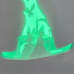 May include: A green, illuminated witch's hat-shaped light. The design includes silhouettes of a castle, birds, and two figures. The layered design creates depth. The overall theme is fantasy and magic, suitable for a themed room.