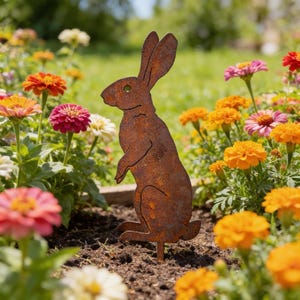 May include: A rusty brown metal rabbit garden stake stands upright in a flower bed. The rabbit has long ears and is surrounded by colorful zinnias and marigolds. The garden art adds a decorative touch to any outdoor space.