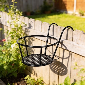 Puede incluir: Cesta colgante de metal negro con diseño de jaula redonda y base tejida. La cesta está suspendida de una valla de madera, con un jardín y vegetación al fondo. Diseñada para sostener una planta en maceta.