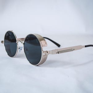 Könnte beinhalten: Steampunk-Sonnenbrille mit runden, dunklen Gläsern und einem silberfarbenen Metallrahmen. Die Brille hat ein einzigartiges Design mit einem Federscharnier an den Bügeln und schwarzen Ohrenstücken. Ein stilvolles Accessoire.