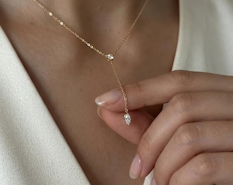 Puede incluir: Un delicado collar de oro con tres piedras preciosas claras y brillantes. El collar presenta un diseño en forma de Y, con una sola piedra en la parte superior y dos piedras en la parte inferior. El collar está siendo sostenido por una persona.