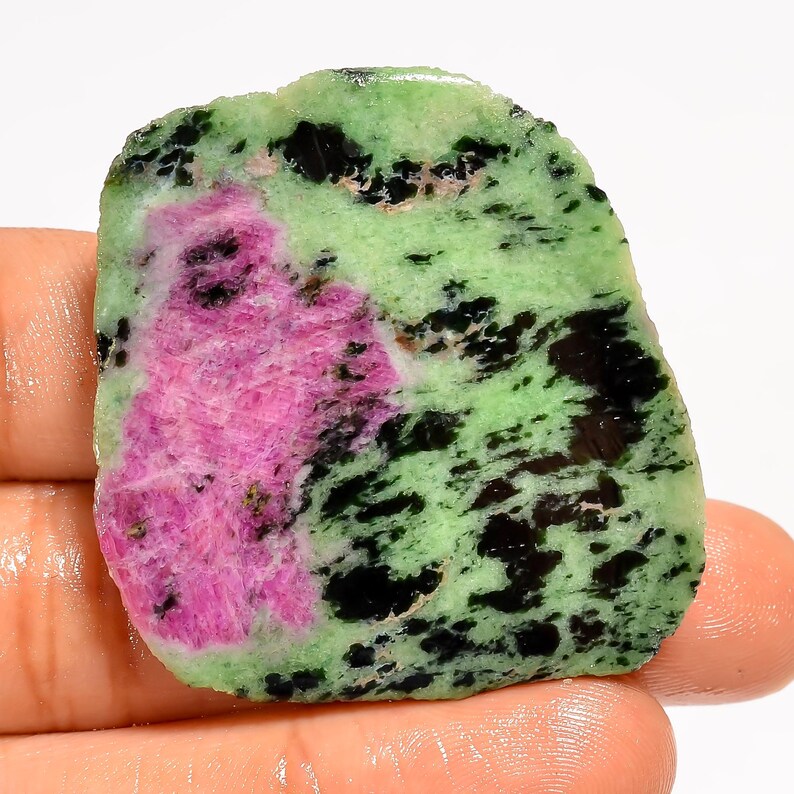 Puede incluir: Una gema en bruto con una vibrante mezcla de colores. La piedra muestra una gran zona de color rosa brillante, rodeada por una base verde claro. Manchas negras oscuras est&aacute;n dispersas por la superficie, creando un patr&oacute;n &uacute;nico. La piedra se sostiene en una mano.