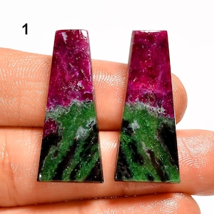 May include: Two rectangular gemstones with a vibrant color scheme. The top portion is a deep magenta, transitioning to a green and black base. The gemstones are held in a hand, showcasing their polished surfaces.