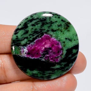 Peut inclure: Une pierre précieuse ronde à la surface polie. La pierre présente un mélange de couleurs vives, notamment du vert, du noir et une tache centrale rose foncé. La pierre est tenue dans une main, mettant en valeur sa taille et sa forme.