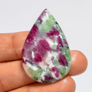 Op de afbeelding: Een gepolijste, druppelvormige robijn-zoisiet edelsteen met een glad, cabochon oppervlak met duidelijke insluitingen van levendige magenta-rode robijnkristallen die contrasteren met een gevlekte achtergrond van ondoorzichtig wit en lichtgroen zoisiet.