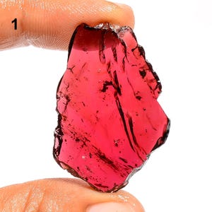 May include: A raw, translucent red garnet gemstone with an irregular, angular shape and a polished, glassy surface texture. The stone displays deep crimson hues and natural internal fractures, showcasing a vibrant, saturated red color throughout.