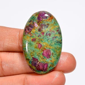 Puede incluir: Gema ovalada con una vibrante mezcla de colores. La piedra presenta una base de tonos verdes y dorados moteados, salpicada de formaciones cristalinas rojas oscuras y moradas. La superficie pulida refleja la luz, resaltando los patrones naturales.