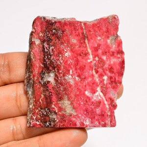 Peut inclure: Spécimen de pierre naturelle brute, de couleur rouge vif, parsemé de veines et de taches blanches et grises. La pierre présente une surface texturée et une forme irrégulière, mettant en valeur son état brut et non poli. La pierre est tenue dans une main.