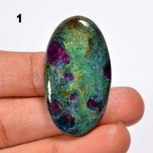 Peut inclure: Une pierre précieuse ovale avec un mélange de couleurs. La pierre présente une base de teintes vertes et bleues, parsemée d'inclusions rouge foncé et violettes. Le coin supérieur gauche porte le chiffre "1" en noir.