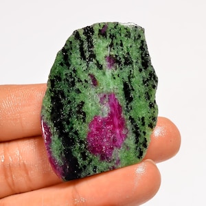Puede incluir: Una gema de zoisita en bruto con una vibrante mezcla de colores. La piedra presenta una base verde con motas negras y un prominente centro de color magenta. La forma irregular y la textura natural resaltan su estado bruto y sin pulir.