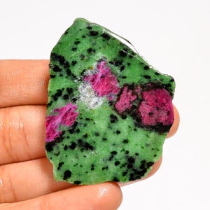 May include: A rough gemstone with a green base, black speckles, and patches of pink ruby. The stone's irregular shape highlights its natural texture and colour variations. The ruby areas have a crystalline appearance, adding to its visual appeal.
