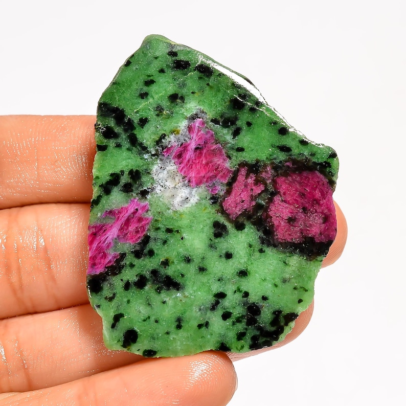 May include: A rough gemstone with a green base, black speckles, and patches of pink ruby. The stone's irregular shape highlights its natural texture and colour variations. The ruby areas have a crystalline appearance, adding to its visual appeal.