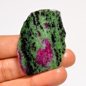 May include: A rough-cut zoisite gemstone with a vibrant mix of colors. The stone features a base of green with black speckles and a prominent magenta-colored center. The irregular shape and natural texture highlight its raw, unpolished state.