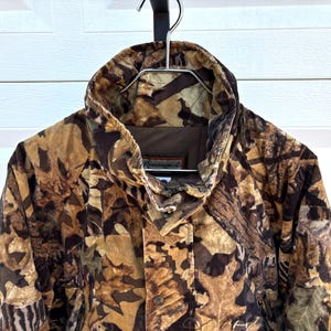 May include: A Remington brand jacket featuring a brown, tan, and black camouflage pattern of leaves and branches. The jacket has a high collar, a front zipper closure, and a brown interior lining. The brand name Remington is visible on the label.