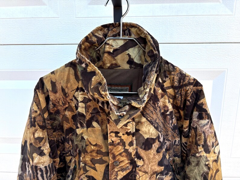 May include: A Remington brand jacket featuring a brown, tan, and black camouflage pattern of leaves and branches. The jacket has a high collar, a front zipper closure, and a brown interior lining. The brand name Remington is visible on the label.