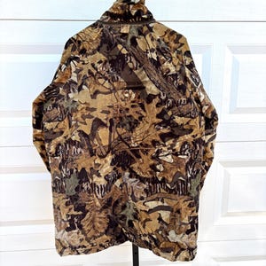 May include: A long-sleeve jacket featuring a detailed camouflage pattern of brown, tan, and dark green autumn leaves and branches. The garment has a high collar, a full-length front zipper, and a textured fabric finish throughout the exterior.