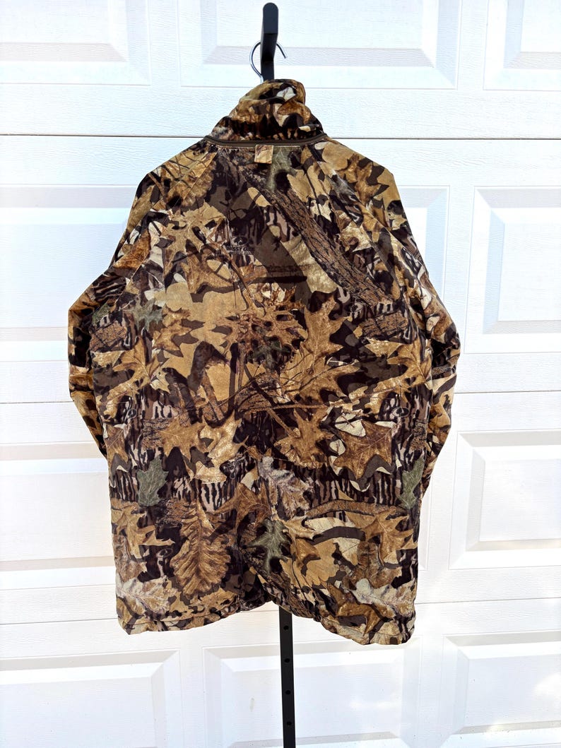 May include: A long-sleeve jacket featuring a detailed camouflage pattern of brown, tan, and dark green autumn leaves and branches. The garment has a high collar, a full-length front zipper, and a textured fabric finish throughout the exterior.