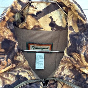May include: A Remington brand jacket featuring a brown and tan camouflage leaf pattern. The interior is lined with solid brown fabric and includes a white tag detailing material composition and size M (38-40). A metal zipper is visible at the center.