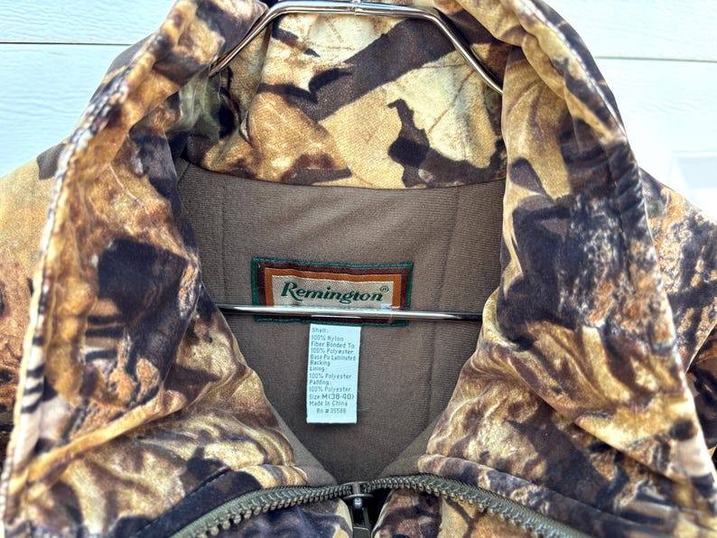 May include: A Remington brand jacket featuring a brown and tan camouflage leaf pattern. The interior is lined with solid brown fabric and includes a white tag detailing material composition and size M (38-40). A metal zipper is visible at the center.