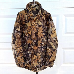 May include: A long-sleeve jacket featuring a brown, tan, and black forest leaf camouflage pattern. The jacket has a high collar, a front zipper closure concealed by a snap-button placket, and two large rectangular front pockets with flaps.