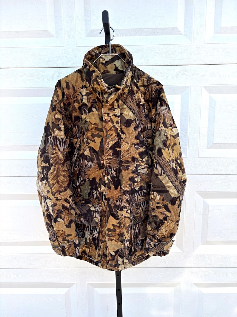 May include: A long-sleeve jacket featuring a brown, tan, and black forest leaf camouflage pattern. The jacket has a high collar, a front zipper closure concealed by a snap-button placket, and two large rectangular front pockets with flaps.
