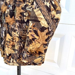 May include: A long-sleeve jacket featuring a camouflage pattern of brown, tan, and black autumn leaves. The jacket includes a front button closure, a collar, and two large rectangular pockets on the lower front with button-down flaps.