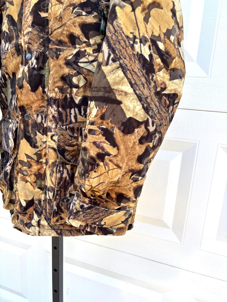 May include: A long-sleeve jacket featuring a camouflage pattern of brown, tan, and black autumn leaves. The jacket includes a front button closure, a collar, and two large rectangular pockets on the lower front with button-down flaps.