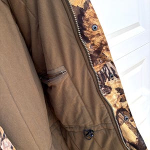 May include: A jacket featuring a brown interior lining with a zippered pocket and a drawstring toggle. The exterior displays a camouflage pattern of brown and tan leaves. A metal zipper runs vertically along the front, secured by black snap buttons.
