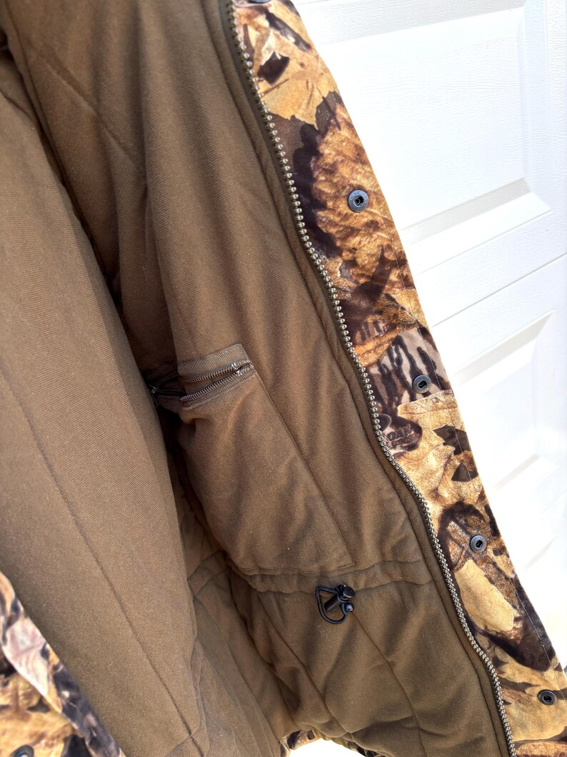 May include: A jacket featuring a brown interior lining with a zippered pocket and a drawstring toggle. The exterior displays a camouflage pattern of brown and tan leaves. A metal zipper runs vertically along the front, secured by black snap buttons.