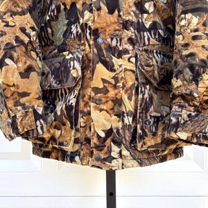 May include: A heavy winter jacket featuring a detailed camouflage pattern of brown, tan, and black autumn leaves. The jacket includes two large front pockets with flaps, a central button-down placket, and adjustable cuffs with hook-and-loop fasteners.
