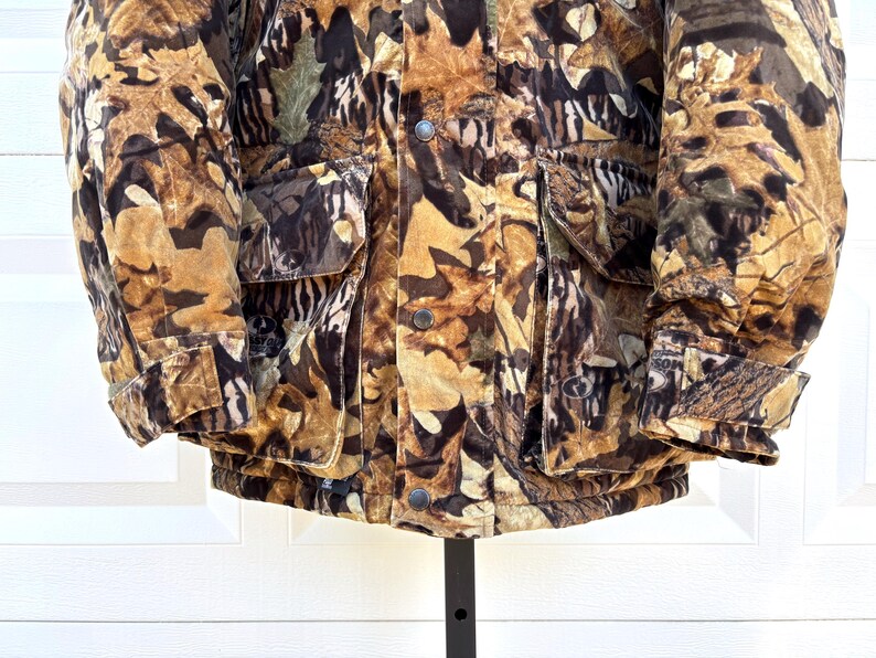 May include: A heavy winter jacket featuring a detailed camouflage pattern of brown, tan, and black autumn leaves. The jacket includes two large front pockets with flaps, a central button-down placket, and adjustable cuffs with hook-and-loop fasteners.