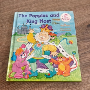Puede incluir: Un libro infantil titulado "The Popples and King Most" con una colorida ilustración de un rey y personajes de dibujos animados. La portada es azul claro con un castillo y un logo de un Popple rosa.