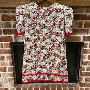 May include: A vintage floral print dress with a white background and a vibrant pattern of red, blue, and yellow flowers. The dress features short sleeves with red trim, a red hemline, and a round neckline. The dress is hanging on a wooden hanger.
