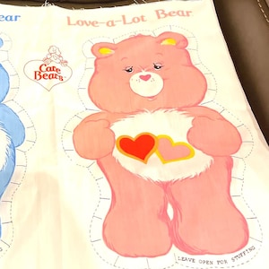 May include: A white fabric panel featuring a printed Love-a-Lot Bear Care Bears character with a pink body, a white belly patch with two hearts, and a dashed cutting line. The text reads Love-a-Lot Bear, Care Bears, and Leave open for stuffing.