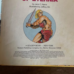K&ouml;nnte beinhalten: Ein Vintage Golden Book mit einer Masters of the Universe-Illustration. Das Cover zeigt einen muskul&ouml;sen, blonden Charakter mit einem Schwert. Der Buchtitel lautet "Of Eternia" von Jack C. Harris, illustriert von Jeffrey Oh.