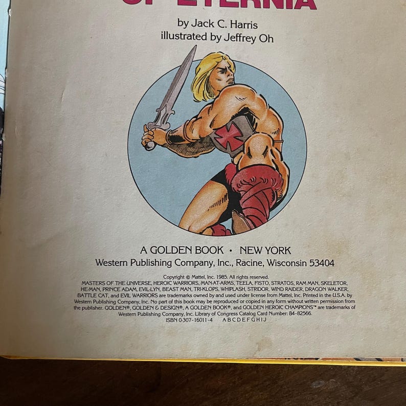 K&ouml;nnte beinhalten: Ein Vintage Golden Book mit einer Masters of the Universe-Illustration. Das Cover zeigt einen muskul&ouml;sen, blonden Charakter mit einem Schwert. Der Buchtitel lautet "Of Eternia" von Jack C. Harris, illustriert von Jeffrey Oh.