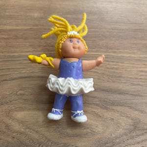 Puede incluir: Una muñeca bailarina vintage de Cabbage Patch Kids. La muñeca tiene cabello de hilo amarillo, ojos azules y lleva un maillot azul con detalles blancos y un tutú blanco. La muñeca mide aproximadamente 13 cm de altura.
