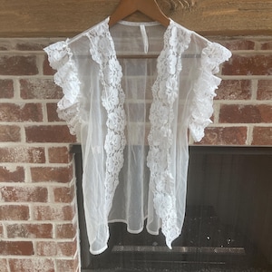 May include: A sheer white lace cover-up with ruffled sleeves hangs on a wooden hanger. The garment features floral lace trim down the front and around the sleeves. The background includes a brick fireplace.