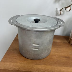 May include: A cylindrical aluminum cooking pot with a matching lid featuring a small black knob. The pot has two curved side handles integrated into the rim and two horizontal embossed lines on the side of the body, showing a metallic finish.