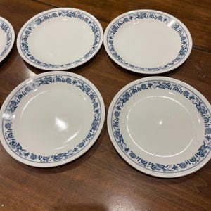 May include: A set of four round white ceramic plates featuring a blue floral pattern along the rim. Each plate has a smooth, glossy finish and a consistent design of interconnected blue flowers and leaves encircling the outer edge of the surface.