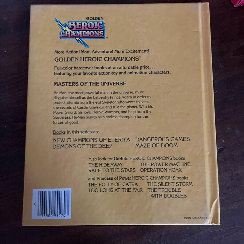 K&ouml;nnte beinhalten: R&uuml;ckseite eines Vintage-Buches von Golden Heroic Champions. Der beige Umschlag zeigt blauen und goldenen Text, darunter den Titel und eine Beschreibung der Masters of the Universe-Serie. Weitere Titel der Serie sind ebenfalls aufgef&uuml;hrt.
