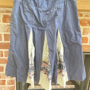 May include: A knee-length blue skirt featuring a series of inverted pleats that reveal cream-coloured fabric panels. The cream panels are decorated with a detailed graphic print of sailing ships in black, orange, and muted red ink lines.