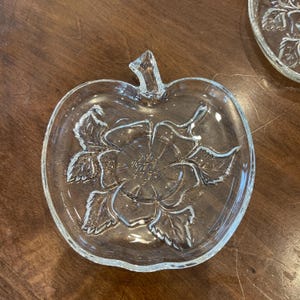 Può includere: Piatto da portata trasparente a forma di mela con un motivo floreale in rilievo. Il piatto presenta un gambo e un motivo dettagliato di fiori e foglie in rilievo. Il vetro trasparente permette di vedere il contenuto, adatto per servire antipasti o dessert.