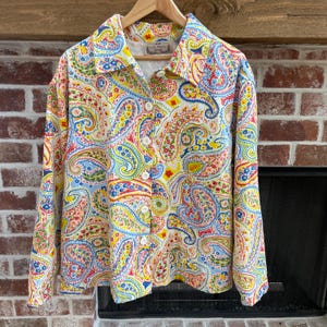 May include: A button-down shirt with a vibrant paisley pattern in red, yellow, blue, and white. The long-sleeved shirt has a collared neckline and white buttons. The shirt is hanging on a wooden hanger.