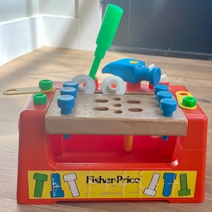 May include: A vintage Fisher-Price toy workbench, model 927, in red with a wooden work surface. Includes a green screwdriver, blue hammer, and various colorful pegs and bolts. The toy has a yellow sticker with the Fisher-Price logo.