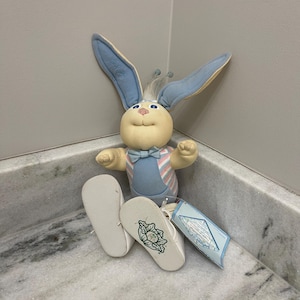 May include: A plush bunny doll with blue and cream colors, wearing a bow tie and striped vest. The bunny has long blue ears and is sitting next to a pair of white baby shoes. A blue tag is attached.