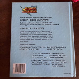 K&ouml;nnte beinhalten: R&uuml;ckseite eines Golden Heroic Champions Buches. Das Buch enth&auml;lt Text &uuml;ber Masters of the Universe und andere Titel der Serie. Das Cover ist hellblau mit einem Logo oben.