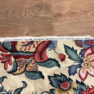 以下が含まれることがあります： 赤、青、緑、タンの色合いの大きな花柄のクリーム色の生地。生地は、様式化された花と葉の繰り返しデザインが特徴です。「GREENFIELD CLASSICS」の文字が上端に印刷されています。